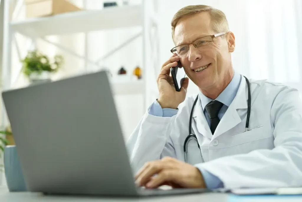 Doctor on a laptop in his office, talking to a patient on the phone.
